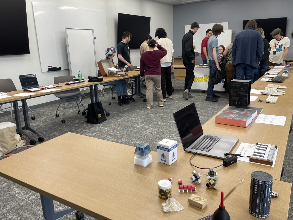 A view of the entire meetup space, showing three flanking tables with keyboards, keyboard accessories and tools, a typewriter, and a book about the history of keyboards. At the end of the room, people gather to see Joe Scotto's handiwork.