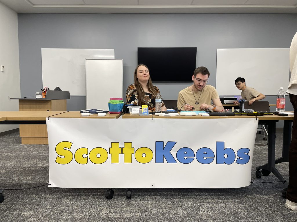 Jordan and Joe of ScottoKeebs at a table featuring a large sign reading "ScottoKeebs" and a sampling of his handwired boards.