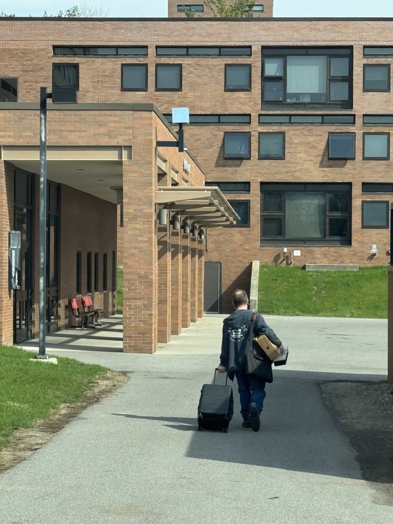 A man walking away from the camera, pulling luggage behind him and carrying a keyboard box and a toolkit.